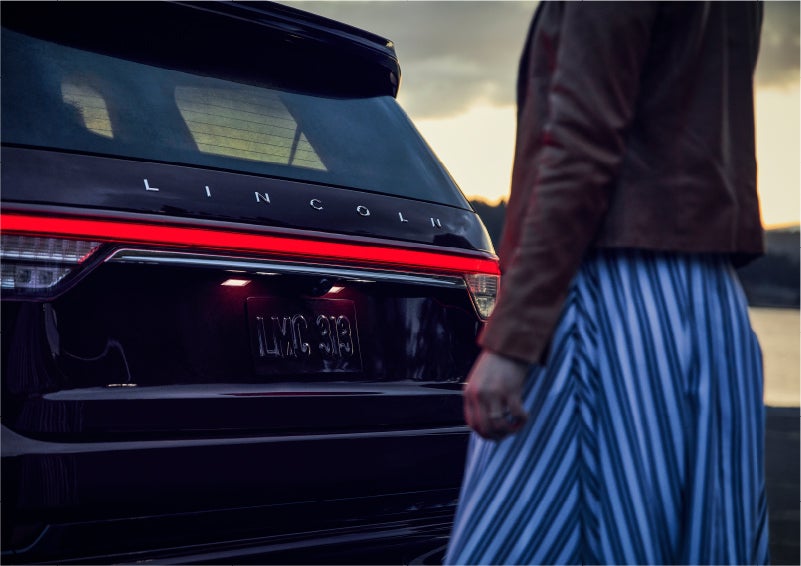 A person is shown near the rear of a 2023 Lincoln Aviator® SUV as the Lincoln Embrace illuminates the rear lights | Koons Lincoln of Bethesda in Silver Spring MD
